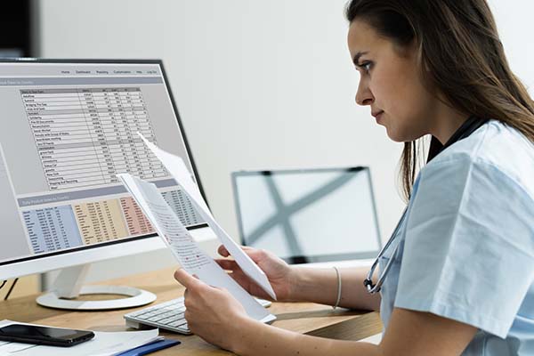 A young female medical professional with a stethoscope around her neck is sitting at a desk, reviewing a paper document while looking at a spreadsheet on a computer screen.