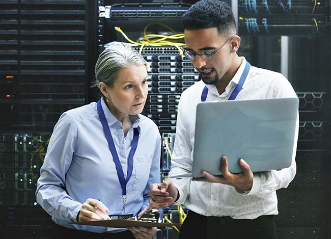 Elevate Technology two IT professionals, a woman and a man, standing in a server room, examining a laptop screen and a clipboard.