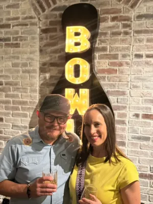 Two colleagues smiling at a social event with drinks near an illuminated sign, representing Elevate Technology and professional technology management for mobile.