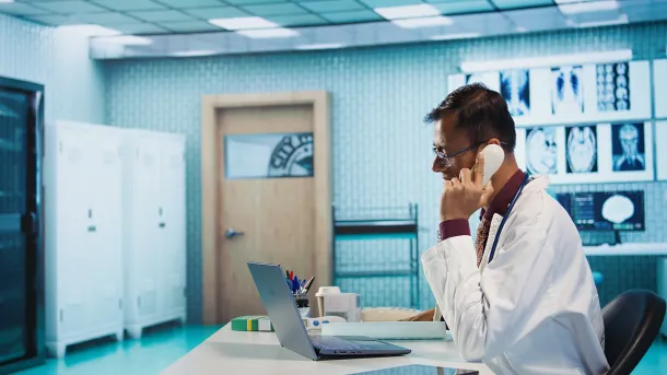 Doctor using a desk phone while sitting at a laptop in a medical office, illustrating how Elevation Technology empowers patient-centered communication in healthcare.